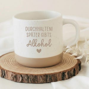 Könnte beinhalten: Eine weiße Keramik-Tasse mit schwarzem Henkel und dem Text "But First, Coffee" in fetter, schwarzer Schrift. Die Tasse steht vor einem verschwommenen Hintergrund, der eine gemütliche Umgebung suggeriert.