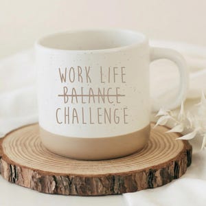 Pode incluir: Uma caneca de cerâmica branca com o texto "WORK LIFE CHALLENGE" em letras castanhas. A palavra "BALANCE" está riscada. A caneca está sobre um descanso de copo redondo de madeira com borda natural.