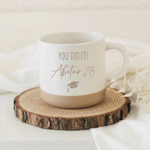Könnte beinhalten: Eine gesprenkelte, weiße Keramik-Tasse mit beigem Boden und Henkel. Die Tasse trägt den Text "YOU DID IT! Abitur 26" mit einer Grafik einer Abschlusskappe. Die Tasse steht auf einer Holzscheibe.