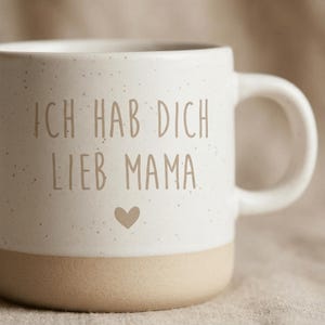 Op de afbeelding: Cr&egrave;mekleurige keramische mok met de Duitse tekst "ICH HAB DICH LIEB MAMA" in bruine letters en een hartje. De onderkant van de mok is lichtbruin en het handvat is wit.