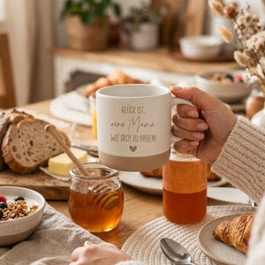 May include: A white ceramic mug with a speckled finish and a tan base is held by a person. The mug has the German text "Gl&uuml;ck ist, eine Mama, wie dich zu haben!" and a heart symbol. The mug is on a table with breakfast items.