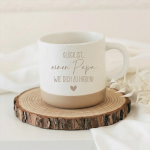 Op de afbeelding: Witte keramische mok met een bruine basis en oor, met de Duitse tekst "Gl&uuml;ck ist, einen Papa, wie dich zu haben!" en een hartje. De mok staat op een houten schijf, perfect als cadeau voor Vaderdag.
