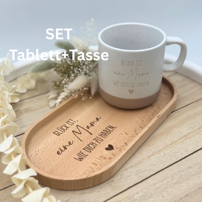 May include: A set including a speckled white mug with a tan base and a wooden serving tray. The mug and tray have German text and a heart symbol. The tray is oval-shaped and made of light-colored wood. The text on the mug and tray reads "Gl&uuml;ck ist eine Mama wie dich zu haben!"
