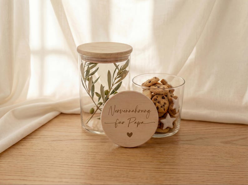 Op de afbeelding: Een heldere glazen pot met een houten deksel en olijftakken erin, naast een kleiner glas gevuld met koekjes. Een houten schijf met de tekst "Nervennahrung f&uuml;r Papa" en een hartje staat ervoor.