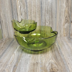 May include: A two-piece green glass serving set. The larger bowl has a wavy rim and sits below a smaller bowl held by a gold-colored metal stand. The set is designed for serving snacks or dips.