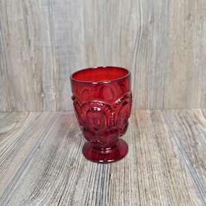 May include: A deep red glass tumbler featuring a raised, repeating circular pattern across its exterior surface. The vessel has a thick, sturdy base and a wide, open rim, showcasing a translucent finish that catches light through its textured design.
