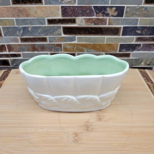 May include: A white ceramic planter with a scalloped rim and leaf design. The interior is a light green colour. The planter is oval-shaped and sits on a wooden surface.