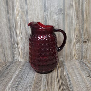 May include: A deep red glass pitcher featuring a prominent hobnail pattern across its rounded body. The vessel includes a curved handle on the side and a wide, moulded pouring spout at the top, showcasing a translucent finish throughout the glass.
