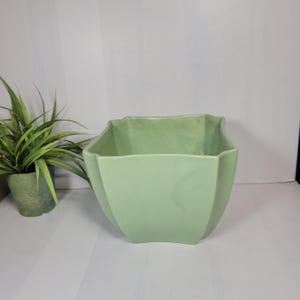 May include: A light green ceramic planter with a geometric design. The planter has a square shape with angled sides and a wavy top edge. A small green potted plant is to the left of the planter.