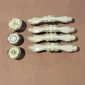 May include: A collection of vintage cabinet hardware, including three white and gold drawer pulls and three decorative knobs. The knobs feature ornate designs, while the pulls have a curved shape. The hardware is likely made of metal.