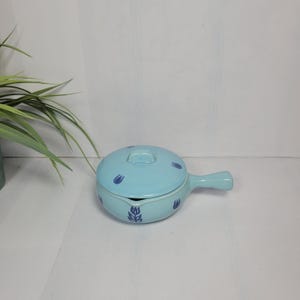 May include: A light blue ceramic saucepan with a matching lid, featuring a dark blue tulip pattern on the exterior and lid. The pot includes a single handle on the side and a small pouring spout, showcasing a smooth, glossy finish throughout.