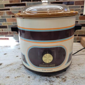 May include: A vintage Rival Crock-Pot slow cooker with a cream-coloured exterior, featuring horizontal brown, orange, and blue stripes. It has a clear glass lid, black side handles, and a central control knob with Off, Low, and High settings.