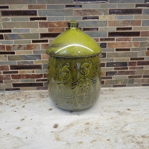 May include: A glossy olive green ceramic canister with a matching lid, featuring a relief design of three stylized figures holding hands, accented with small star motifs and geometric patterns around the base of the figures.