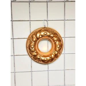 May include: A vintage copper bundt cake pan with a decorative floral design. The pan has a circular shape with a central hole and is suspended from a metal grid. The copper has a warm, reflective finish, suggesting it is well-used.
