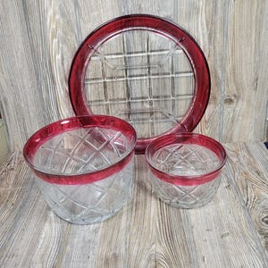 May include: A set of clear glass tableware featuring a deep red rim and a diamond-cut pattern. The collection includes one large circular serving platter, one medium-sized bowl, and one smaller bowl, all sharing a consistent design aesthetic.