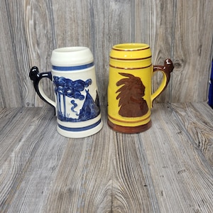 May include: Two ceramic beer steins. The left stein is cream-colored with blue painted illustrations of trees and a teepee, featuring a dark handle. The right stein is yellow with a brown embossed profile of a person wearing a headdress.