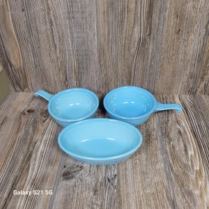 May include: Three light blue ceramic dishes, including two round bowls with side handles and one oval bowl. Each piece features a subtle ribbed texture along the exterior base, providing a consistent look across the set of kitchenware items.