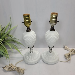 May include: A pair of white milk glass table lamps featuring a hobnail textured bulbous body and a fluted circular base. Each lamp has a metallic gold-toned socket at the top and includes a white electrical cord with a standard two-prong plug.