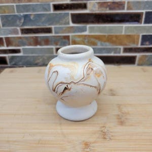May include: A small, round ceramic vase with a matte white base and a swirling marbled pattern in shades of tan, brown, and cream. The vase features a narrow neck, a wide, rounded body, and a solid, circular pedestal base for stability.