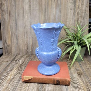 May include: A light blue ceramic vase with a fluted rim and decorative handles. The vase has a textured surface and is sitting on a red book. The vase is approximately 20 cm tall.