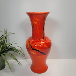 May include: A tall, glossy ceramic vase featuring a vibrant orange base color with swirling patterns of red and yellow highlights. The surface is accented by thin, dark black lines that create a marbled effect across the rounded body and neck.