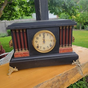 May include: A rectangular mantel clock with a matte black finish, featuring a central circular clock face with black Arabic numerals and gold hands. The clock is accented by vertical red columns with gold-toned capitals and ornate gold feet.