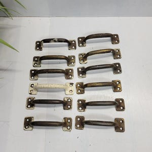 May include: A collection of twelve vintage metal drawer pulls. The pulls have a curved handle design with two mounting plates on each end. The metal has a distressed, aged appearance with a mix of dark brown and off-white paint.