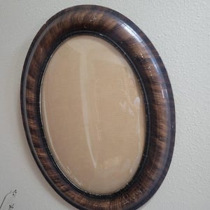 May include: An oval picture frame with a dark brown, faux-wood grain finish and a glossy surface. The frame features a convex glass pane that protrudes outward, creating a rounded, bubble-like appearance within the dark, polished border.