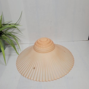 May include: A peach-colored, vintage-style lampshade with a scalloped edge and a ribbed, conical top. The shade has a wide, circular design with vertical ridges radiating from the center. The overall shape is reminiscent of a seashell.