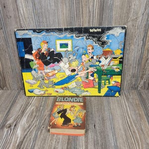 May include: A rectangular jigsaw puzzle featuring a colorful Blondie comic strip scene with characters in a kitchen, accompanied by a small hardcover book titled Blondie No Dull Moments, which depicts a cartoon couple and a child on its front cover.