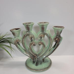 May include: A ceramic candelabra featuring four candle holders arranged in a symmetrical, heart-inspired design. The piece is finished in a glossy light green glaze with dark brown, mottled accents highlighting the curved, scroll-like details.