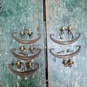May include: A collection of antique drawer pulls and hardware, arranged on a weathered, teal-painted wooden surface. The pulls are a curved, ornate design in a bronze finish, accompanied by brass-colored knobs and screws. Ideal for vintage furniture restoration.