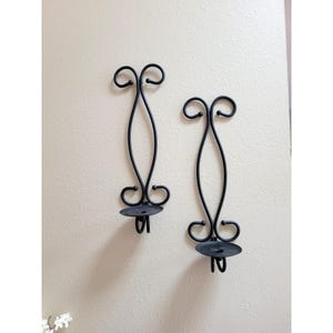 May include: Two black metal wall sconces featuring a vertical, elongated scroll design with a circular base at the bottom for holding a candle. The metal frames are constructed from thin, curved rods that form a symmetrical, open-work pattern.