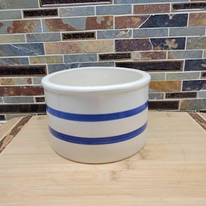 May include: A cylindrical ceramic crock with a smooth off-white finish and two horizontal navy blue stripes encircling the exterior. The vessel features a wide, open top with a thick, rounded rim and a flat base, presenting a simple, functional design.