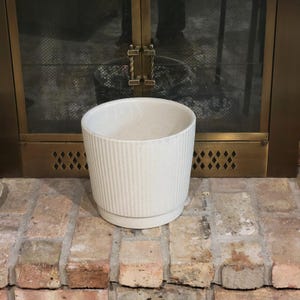 May include: A cream-coloured, ribbed ceramic planter with a cylindrical shape. The planter sits on a matching base and is designed for indoor or outdoor use. The planter is placed on a brick surface.