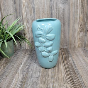May include: A light blue ceramic vase with a smooth, glossy finish and a raised, three-dimensional floral relief design on the front. The vase has a wide, open mouth and a tapered cylindrical shape, showcasing a soft, solid color throughout.