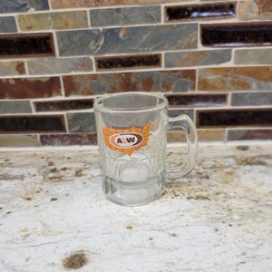 May include: A clear glass mug with a handle and a textured exterior. The mug features an orange and white A&W logo in the shape of the United States. The mug is empty and sits on a white and brown speckled surface.