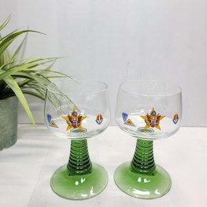 May include: Two clear wine glasses with green stems and bases. Each glass features a decorative emblem with a star and the text "KEEPERS OF THE PEACE". The stems are ribbed, and the bases are circular.