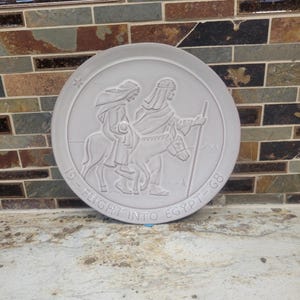 May include: A circular light grey ceramic plate featuring a raised relief design of two figures travelling on a donkey. The text "19 - FLIGHT INTO EGYPT - 68" is embossed along the bottom edge, and a small star is positioned near the top left rim.