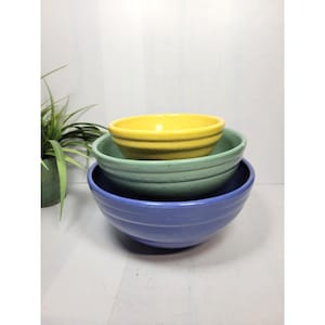 May include: Three ceramic mixing bowls in a nested arrangement. The bowls are in shades of yellow, green, and blue. The yellow bowl sits atop a green bowl, which is above a blue bowl. Each bowl features a ribbed design.