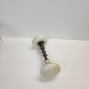 May include: A vintage door knob set featuring two round, off-white porcelain knobs connected by a central metal spindle. The metal components show visible signs of oxidation and wear, with a coiled spring mechanism positioned along the shaft.