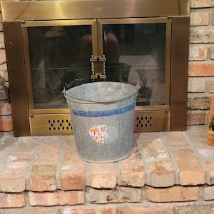 May include: A galvanized metal bucket with a blue stripe and a sticker that says "MA". The bucket is sitting on a brick hearth in front of a fireplace with a brass frame and glass doors.