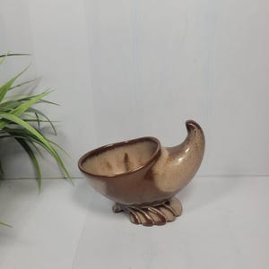 May include: A ceramic cornucopia-shaped planter featuring a two-tone glaze with a dark brown base and rim transitioning into a speckled beige center. The vessel rests on a flared, leaf-like pedestal base with a curved, pointed end.