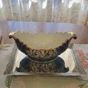 May include: A deep, oval-shaped ceramic bowl with a dark blue exterior featuring a cream-colored floral pattern and a scalloped rim accented with gold trim. The bowl is positioned on a rectangular, silver-toned mirrored tray with an ornate, filigree-style border.