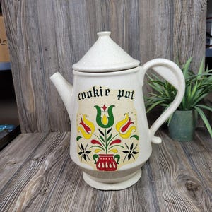 May include: A speckled cream-colored ceramic cookie pot with a conical lid and a handle. The front features a decorative floral design in red, yellow, green, and black, with the text cookie pot printed in black lettering above the illustration.