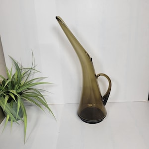May include: A tall, translucent smoke-colored glass pitcher with a wide, conical base that tapers into a long, slender, upward-curving spout. The vessel features a curved glass handle attached to the side, providing a sleek, elongated silhouette.