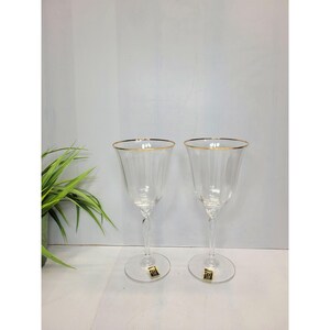 May include: Two clear wine glasses with gold rims. The glasses have a ribbed design and a twisted stem. Each glass has a round base and a small gold label. The glasses are displayed on a white surface.