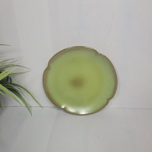 May include: A round, light green ceramic plate with a scalloped edge. The center of the plate has a darker green hue, and the rim is a darker brown. The plate is set against a white background.