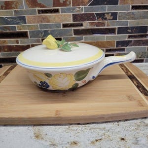 May include: A white ceramic casserole dish with a lid, featuring a yellow rose and blue floral design. The lid is topped with a yellow rose and green leaves. The dish has a handle and a yellow and blue trim. It sits on a wooden cutting board.