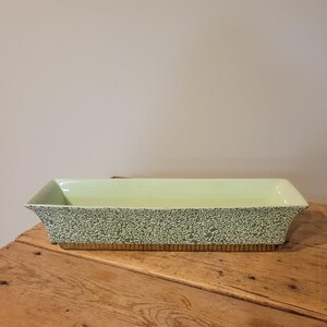 May include: A rectangular ceramic planter with a light green interior and a speckled green and white exterior pattern. The base of the planter features a textured, ribbed gold-toned band that runs along the bottom edge of the vessel.
